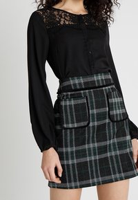 Blusa nera con yolk in pizzo e chiusura a bottone, abbinata a una gonna a quadri con tonalità verde, grigio e nero, dotata di tasche frontali.