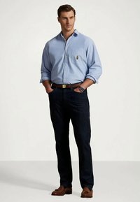 Camisa azul claro com botões e bolso com emblema, jeans escuro, cinto castanho e sapatos castanhos, usados por um homem numa postura neutra.