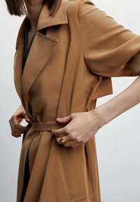 Tan trench coat with a belted waist, short sleeves, and collar. Smooth fabric and notable stitching details at the seams.
