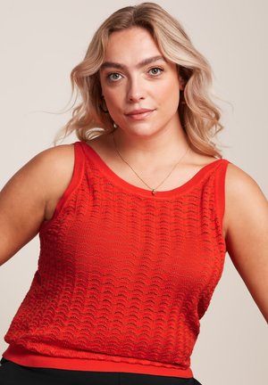 Blonde vrouw met los golvend haar, gekleed in een mouwloze rode textuurtop en een fijn ketting tegen een neutrale achtergrond.