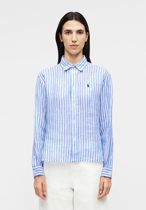 Femme portant une chemise boutonnée à manches longues à rayures verticales bleues et blanches, avec un pantalon blanc, debout devant un fond uni.