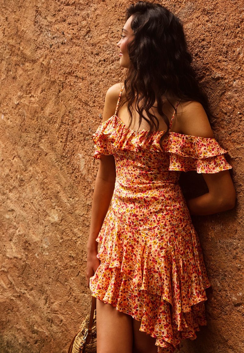 Femme aux longs cheveux foncés portant une robe orange fleurie hors épaules, appuyée contre un mur brun texturé, tenant un sac tressé.
