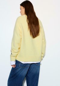 Żółty sweter z dzianiny o miękkiej fakturze, luźnym fasonie i prążkowanym wykończeniu; noszony na białej koszuli i zestawiony z niebieskimi dżinsami.