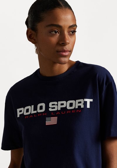 T-shirt en coton bleu marine avec "POLO SPORT" en lettres blanches en gras, accentué de rouge et avec un petit graphique du drapeau américain. Manches courtes et col rond.