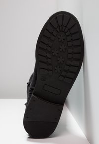 Czarny spód buta z teksturowanej gumowej powierzchni, z bieżnikiem o różnych kształtach oraz logo producenta z boku.