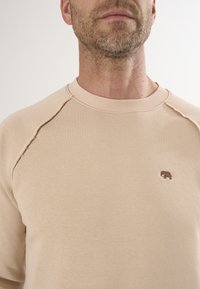 Beige Sweatshirt mit geripptem Ausschnitt, ausgestattet mit einem kleinen braunen Elefantenlogo auf der linken Brust und rohen Kanten an den Schultern.