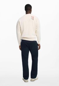 Világos krémszínű pulóver, lekerekített nyakkivágással, bordázott mandzsettákkal és szegéllyel. A hátán nyomott minta található. Tengerészkék nadrággal párosítva.