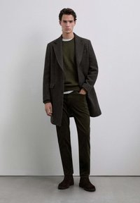 Abrigo de lana gris oscuro con solapas de muesca, suéter de punto verde, pantalones de pana verde oliva y botines de ante marrón. Diseño minimalista.