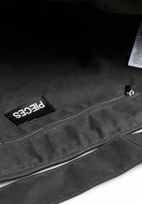 Juodos drobės maišo vidus su matomu užtrauktuku ir etikete su užrašu "PIECES". Strukturinė medžiaga su matinio paviršiaus apdaila.