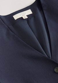 Veste en lin bleu marine avec col cranté, fermeture à un bouton, tissu lisse et texturé avec un éclat subtil.