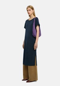 Długa granatowa sukienka tunika z krótkimi rękawami, z bocznymi rozcięciami, w połączeniu z szerokimi beżowymi spodniami i fakturowaną fioletową torbą na ramię.