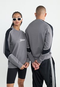 Grå hettegenser med svarte detaljer og hvite kanter, med en logo. Modellen har på seg svarte shorts og holder hendene sammen. En annen modell står bak.