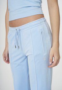 Ljusblåa velourmjukisbyxor med elastisk midjeresår, dragsko, sidofickor med dragkedja samt broderad logotypdetalj på vänster ben.