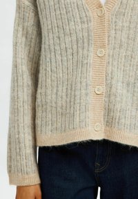 Beige ribbattu cardigan V-kaulusega, millel on helebeez nööbivalik ja varrukad, valminud tekstuuriga kootud kangast, kombineeritud tumedate teksadega.