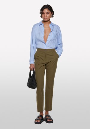 Femme portant une chemise boutonnée bleu clair, un pantalon vert olive, des sandales noires, tenant un sac à main tissé noir, se tenant devant un fond uni.