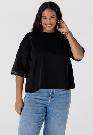 Femme souriante avec de longs cheveux noirs portant un haut noir ample avec des manches en dentelle et un jean bleu clair, posant avec une main près de son oreille.