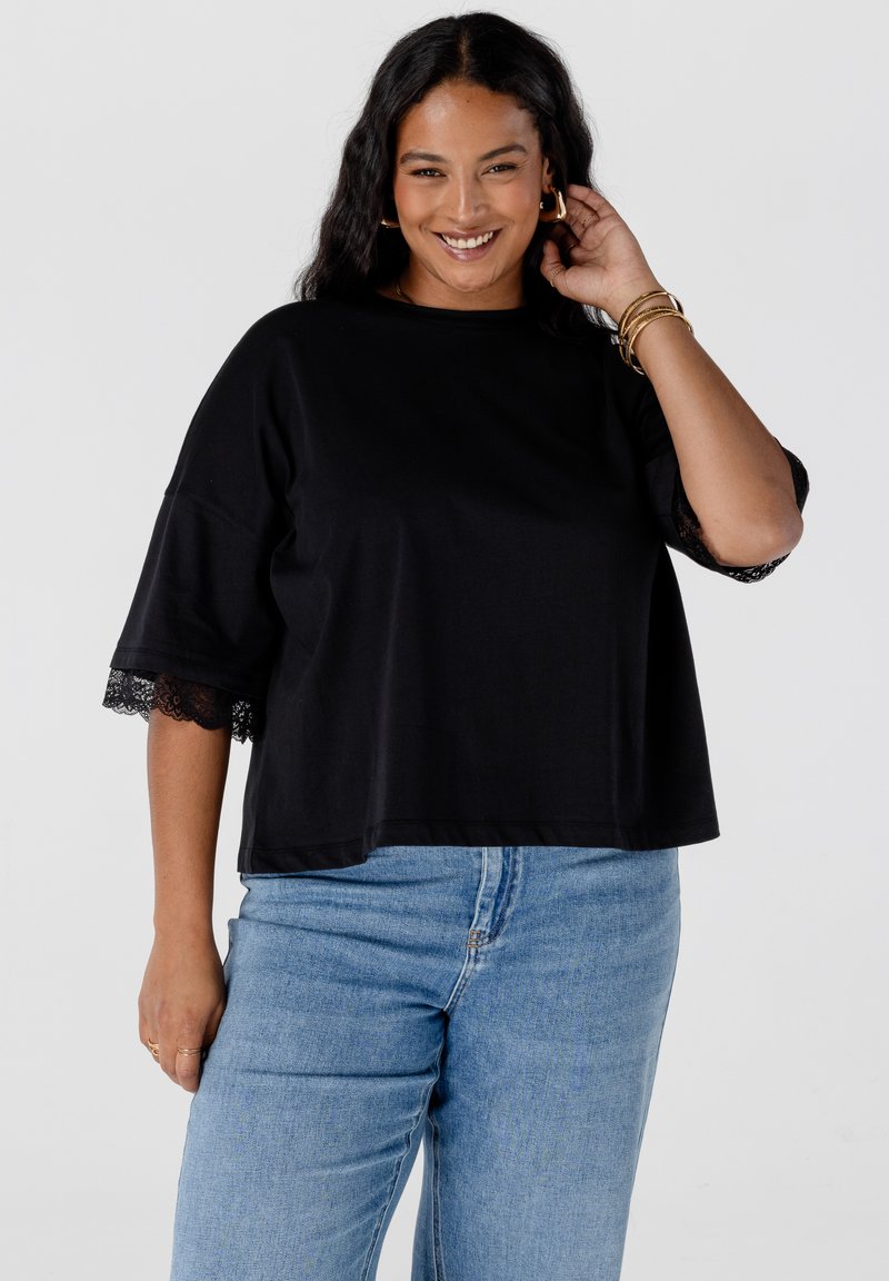 Glukkig glimlachende vrouw met lang donker haar, gekleed in een zwarte losse top met kantmouwen en lichtblauwe jeans, poseert met haar hand bij haar oor.