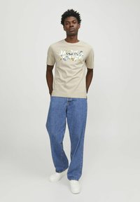 Beige t-skjorte med blomsterdesign og teksten "JACK & JONES," paret med løse, mørkeblå jeans og hvite sneakers. Glatt stoff, avslappet passform.