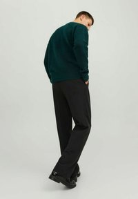Pull en tricot vert foncé avec un motif texturé, associé à un pantalon noir et des chaussures noires. La personne pose avec les mains dans les poches.