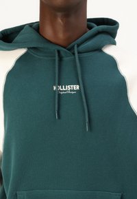 Hooded sweatshirt in dark teal with white raglan sleeves. Features a front pocket and embroidered "HOLLISTER" logo. Soft texture.