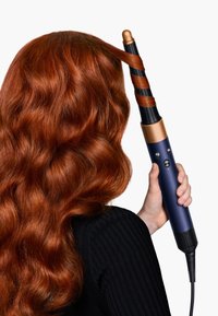 Piastra arricciacapelli con un corpo blu navy, accenti dorati e un cilindro a motivo spirale. Impugnatura texturizzata, cavo per la connessione elettrica, capelli rossi ondulati visibili.