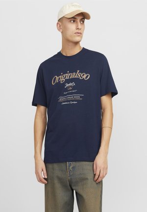 Marineblå bomulds t-shirt med fronttryk i beige. Kombinationen med lyse jeans giver et afslappet og enkelt look. En cremefarvet baseballcap på hovedet.