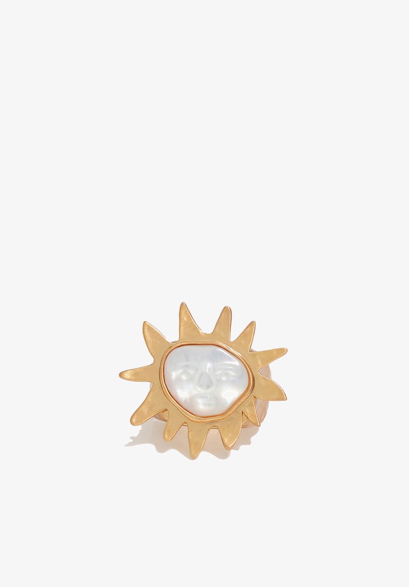 Gold sun-shaped ring with a white pearl center carved into a face, casting a soft shadow on a white background.
