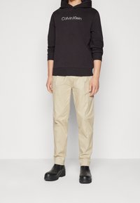 Sweatshirt à capuche noire avec le logo "Calvin Klein", associé à un pantalon beige et des bottes en cuir noir à la cheville, se tenant sur un fond uni.