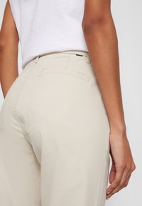 Pantalon beige à texture lisse, avec une poche arrière. La ceinture comprend des passants pour ceinture et une petite étiquette logo est visible.