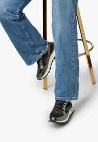 Svarta och gröna sneakers med glansiga detaljer, med en texturerad yta och tjock vit sula, bärs med blå denimjeans.