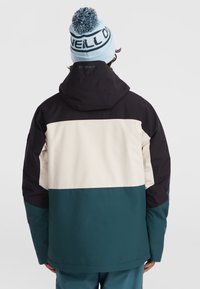 Kleurgeblokte jas met zwarte, crèmekleurige en teal secties; beschikt over een capuchon en sportief ontwerp. Blauwe gebreide beanie met pompon detail.