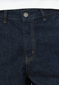 Jean en denim bleu foncé avec surpiqûres jaunes, fermeture à bouton métallique et design à cinq poches. Texture lisse et coupe droite classique.