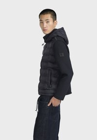 Gilet imbottito nero con cappuccio, caratterizzato da un design trapuntato e tasche con cerniera, abbinato a una maglietta nera a maniche lunghe e jeans in denim.