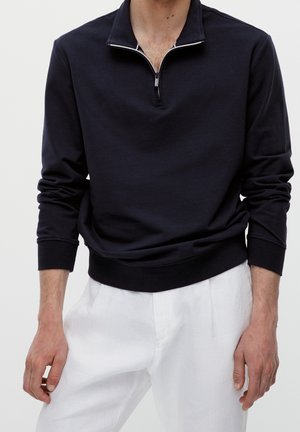 Homme portant un pull noir à manches longues avec une courte fermeture éclair et un pantalon blanc, debout devant un fond uni.