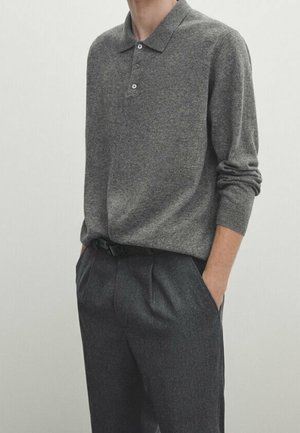 Mężczyzna w szarej, długosplecionej koszulce polo z guzikiem, włożonej do ciemnoszarych spodni z kantem, z rękami w kieszeniach.