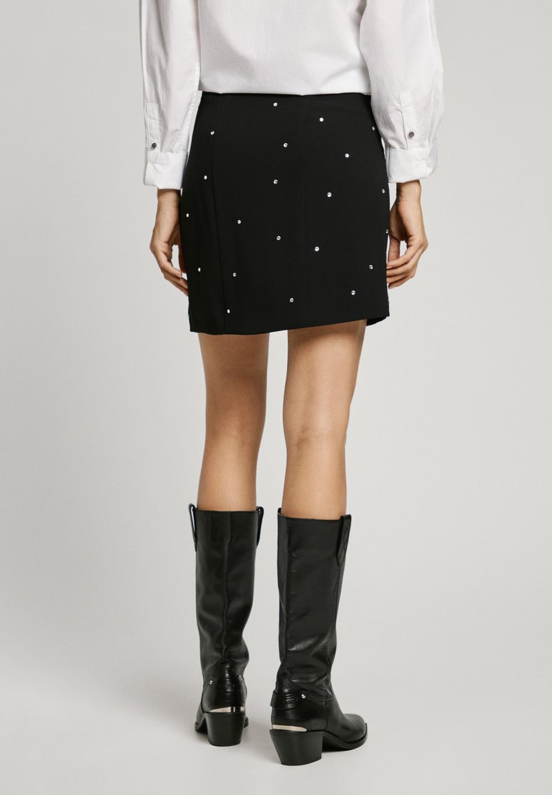 Jupe noire avec de petites strass décoratifs, tissu lisse, longueur au-dessus du genou, associée à des bottes en cuir noir hautes.