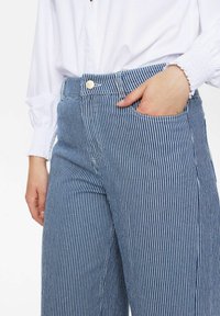 Pantalons larges rayés bleus et blancs avec taille haute, poches latérales et fermeture à bouton doré. Tissu en coton lisse avec des lignes verticales.
