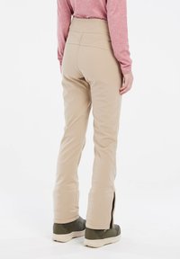 Beige Skihosen mit hohem Bund, schmaler Passform und Reißverschluss an den Knöcheln, kombiniert mit dunkelgrünen Schuhen. Glatte Stofftextur.