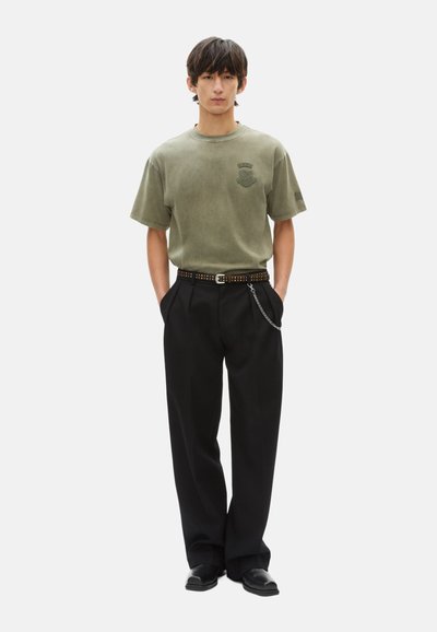 Camiseta de algodón color verde oliva con un logo, combinada con pantalones anchos negros y un cinturón con tachuelas. Unos sencillos zapatos negros completan el look.