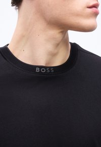 Gros plan sur le cou et la mâchoire d'une personne portant un t-shirt à col rond noir avec "BOSS" imprimé sur le col.
