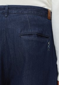 Σκούρο μπλε τζιν πίσω τσέπη με τρεις μικρές κεντητές ραφές σε σχήμα X σε μπεζ, μπλε και σκούρο γκρι χρώματα.