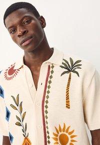 Kortærmet, cremefarvet strikket polo med farverige tropiske og blomsteragtige mønstre, der har knapper og en krave.