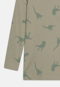 Chemise à manches longues en tissu côtelé kaki avec des silhouettes de dinosaures et de lions verts dans diverses poses, avec des ourlets et des poignets cousus.