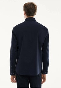 Chemise à manches longues bleu marine avec une texture lisse, col pointu et poignets à boutons ; présente un dos droit et un style minimaliste.