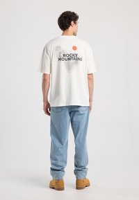 Kerman värinen puuvillainen t-paita lyhyillä hihoilla, jossa on graafinen printti aaltoilevista viivoista ja teksti "ROCKY MOUNTAINS" takana.