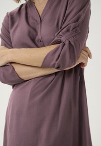 Blouse violette avec un col à encoches, manches retroussées maintenues par un bouton, fabriquée en tissu léger avec une texture lisse.