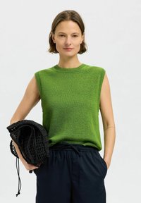 Grünes gestricktes ärmelloses Oberteil mit runder Ausschnittlinie, getragen mit schwarzen Hosen mit Kordelzug und einem schwarzen gehäkelten Beutel. Schlichte Textur.