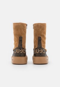 Brązowe botki ze skóry zamszowej z czarnym wzorzystym obcasem, złotymi elementami ozdobnymi i wyrazistym logo z tyłu. Zamek błyskawiczny po bokach.