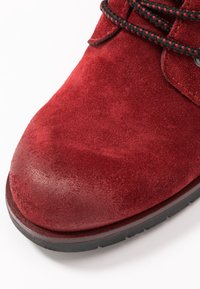 Rote Wildleder-Ankelstiefel mit abgerundetem Zehenbereich, kontrastierenden schwarzen Schnürsenkeln und einer strukturierten Oberfläche. Die Sohle ist schwarz und verfügt über ein Profilmuster für besseren Halt.