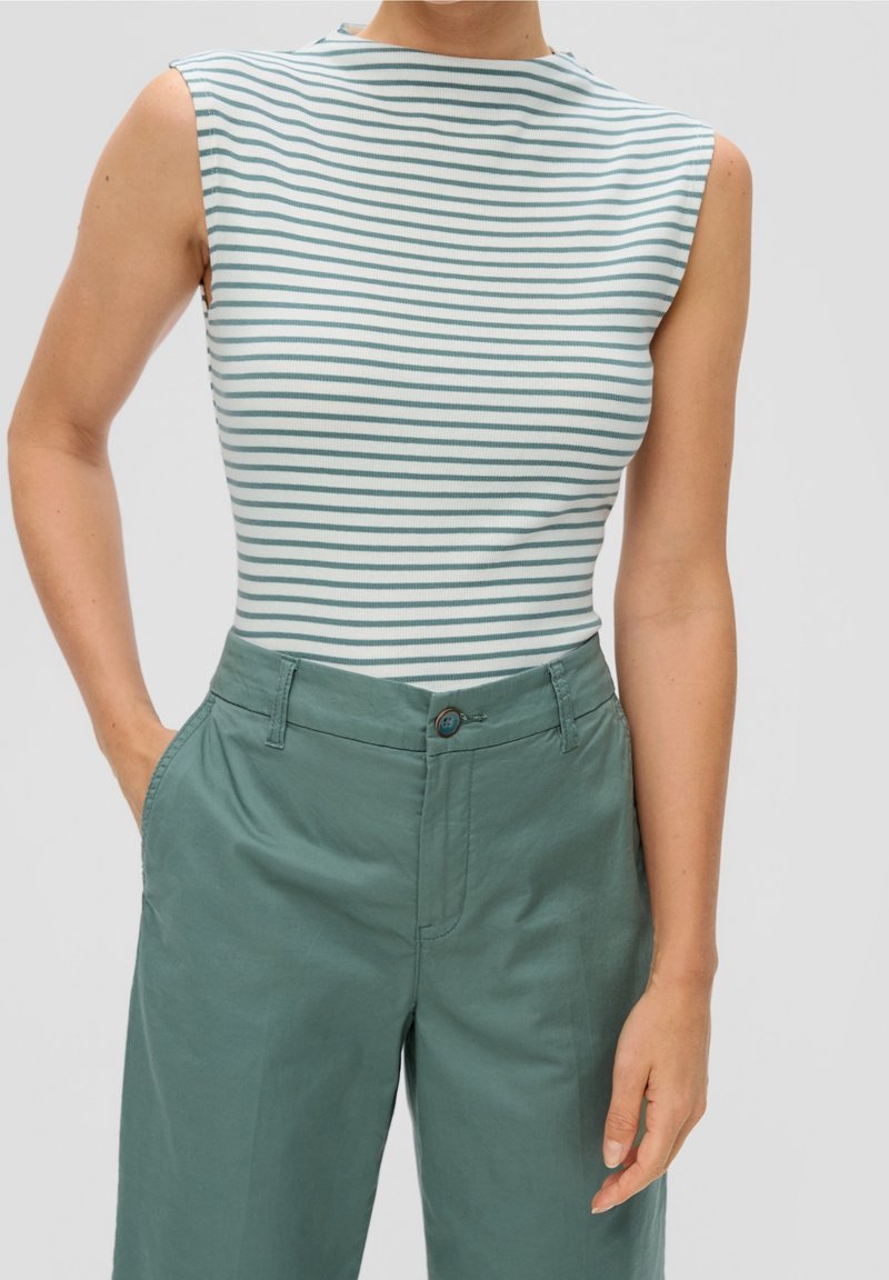 Haut blanc sans manches avec fines rayures horizontales vert foncé, associé à un pantalon ample taille haute vert foncé, mains dans les poches.